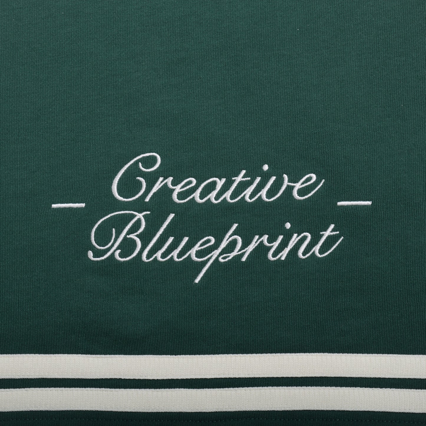 An up-close view of the cream "Creative Blueprint" embroidery design on the bottom of the back of the shirt. "Creative" is stacked on top of "Blueprint". The words are embroidered in a script font and there are two dashes on either side of the embroidery.