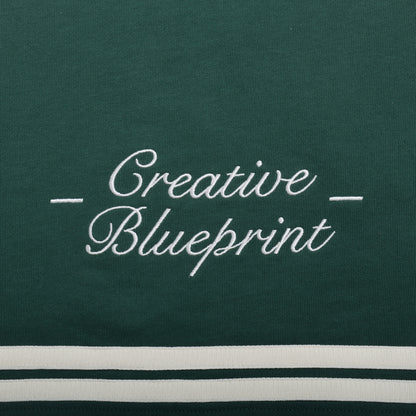 An up-close view of the cream "Creative Blueprint" embroidery design on the bottom of the back of the shirt. "Creative" is stacked on top of "Blueprint". The words are embroidered in a script font and there are two dashes on either side of the embroidery.