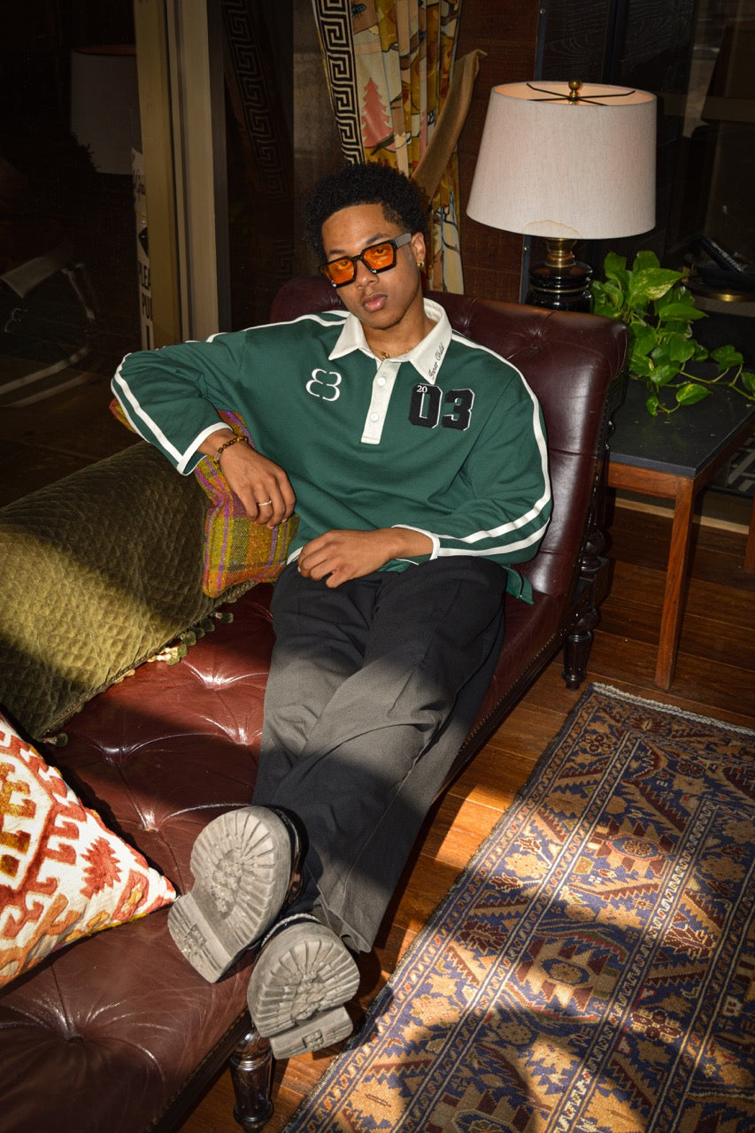 Our model, Darnell, standing 6'0", in our Inner Child Rugby Polo and black slacks, sitting on a brown leather couch with his legs extended, facing the frame.