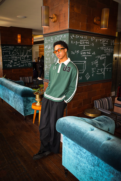 Our model, Darnell, standing 6'0", in our Inner Child Rugby Polo and black slacks, positioned leaning backwards against a chalkboard pillar.